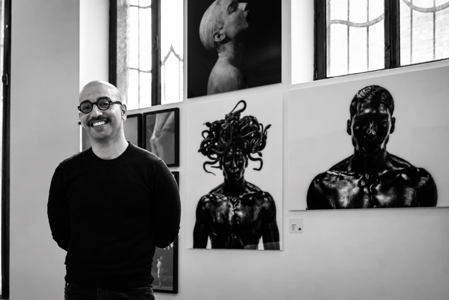 Rafael Montalvo smiles in a gallery beside large black-and-white prints, including a male Medusa crowned with snakes.